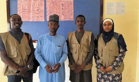 ZAIDF team members and a school leader pose in front of a classroom notice board