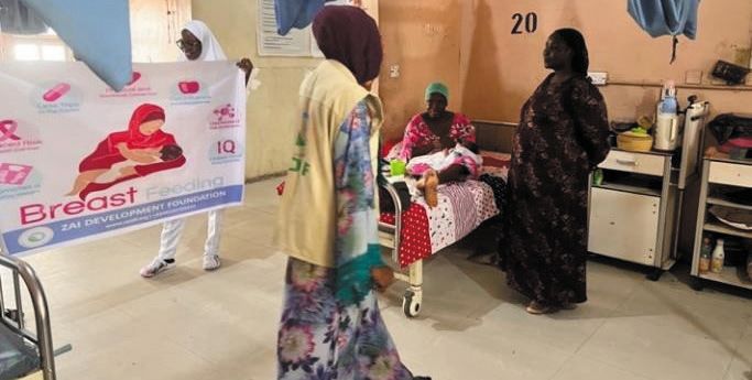 A ZAIDF team member with a breastfeeding banner speaks with a mother at a hospital bedside