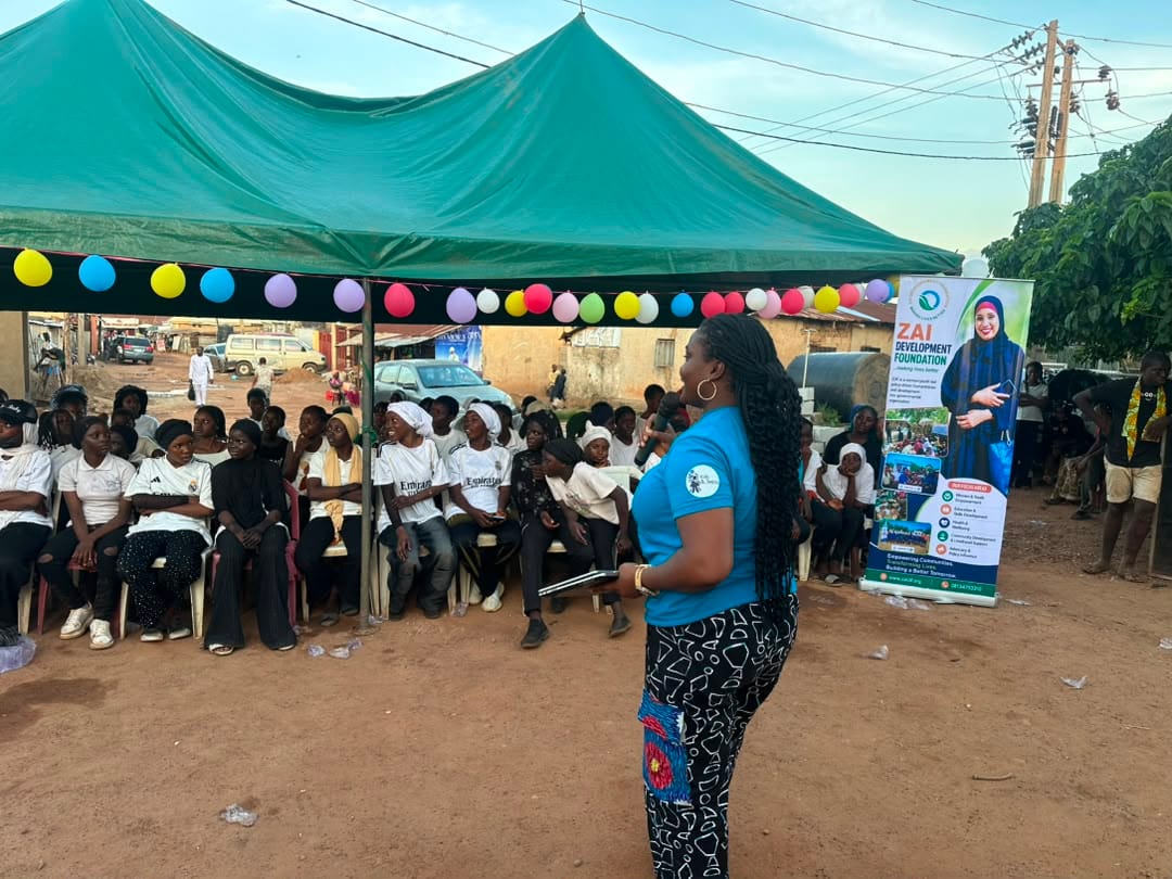 A ZAIDF facilitator in blue addresses a seated audience under green canopies